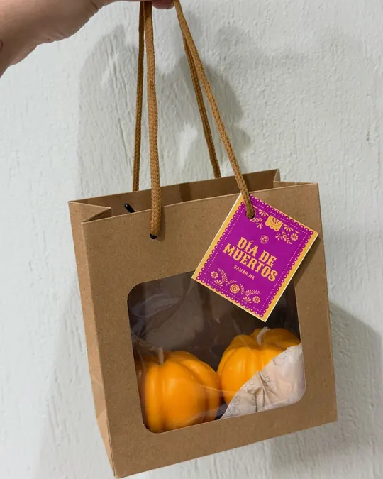 Mano sosteniendo una bolsa kraft de Samar.mx con ventana, calabazas de cera dentro y tarjeta de Día de Muertos