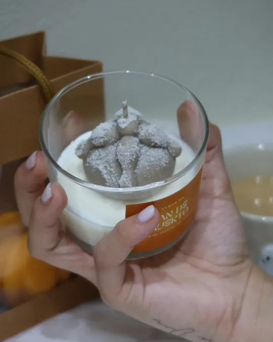 Mano sosteniendo la vela Pan de muerto de Samar.mx, un vaso de cera blanca con un pan de muerto de cera gris encima