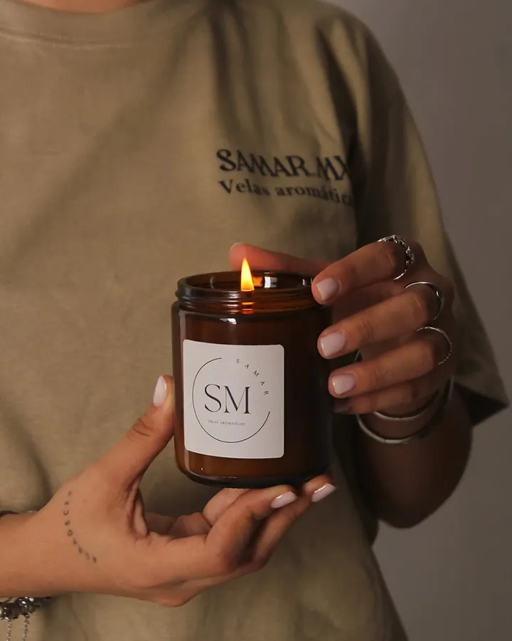Manos sosteniendo una vela encendida de Samar.mx en vaso ámbar con la etiqueta SM